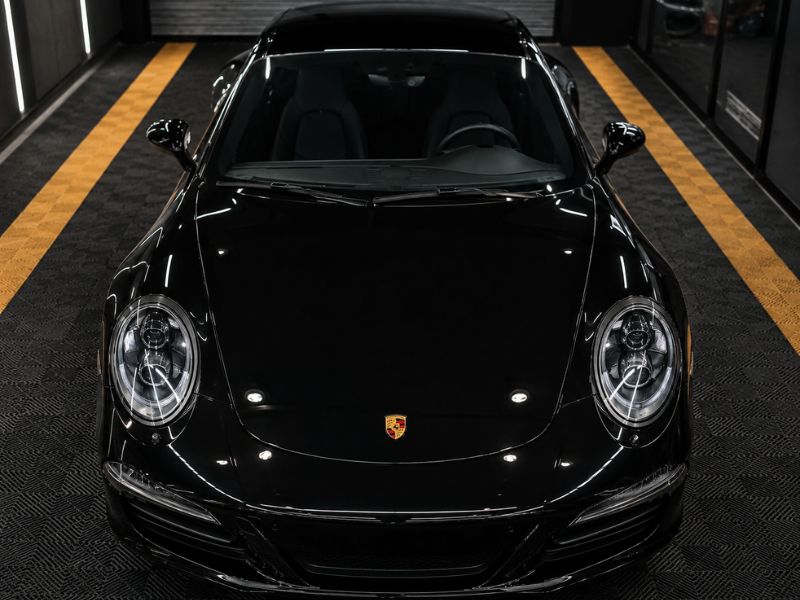 Porsche 911 preto brilhante visto de frente, dentro de garagem sofisticada com piso emborrachado e faixas amarelas, iluminação de teto, acabamento impecável de pintura preta refletindo luzes, fotografia automotiva realista de alto contraste