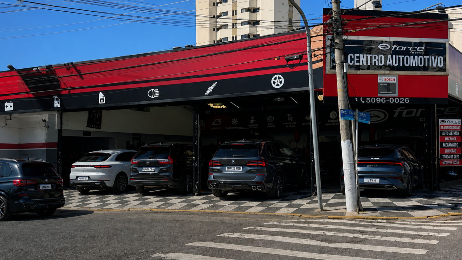 Fachada do centro automotivo Force Car Service com toldo vermelho e preto, letreiro 'Centro Automotivo', veículos estacionados na entrada da oficina, prédios residenciais ao fundo, faixa de pedestre em primeiro plano, Campo Belo, São Paulo, fotografia realista diurna